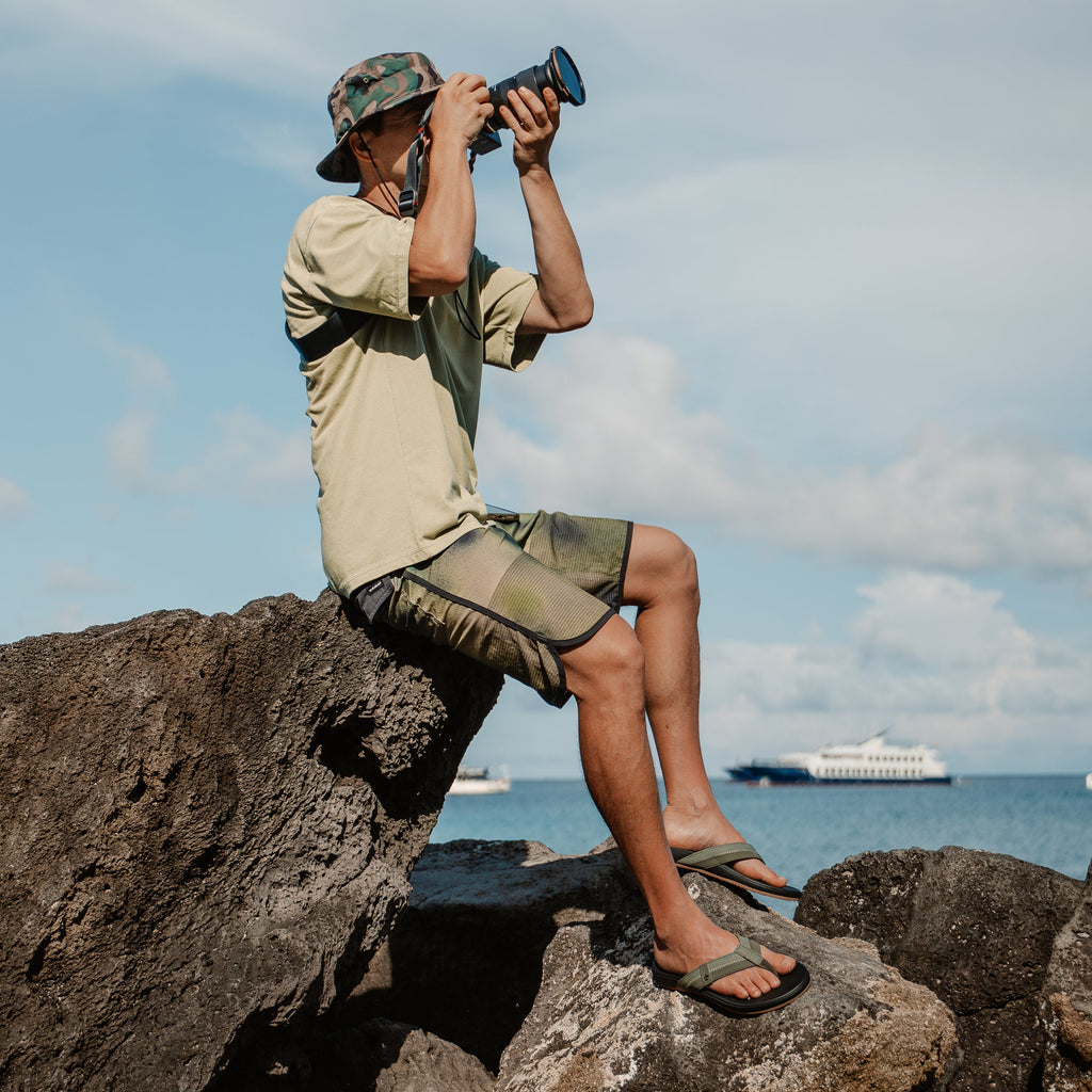 Adult man wearing Cushion Phantom 2.0 sandals facing right, seated on rocks by the ocean with camera in hand.