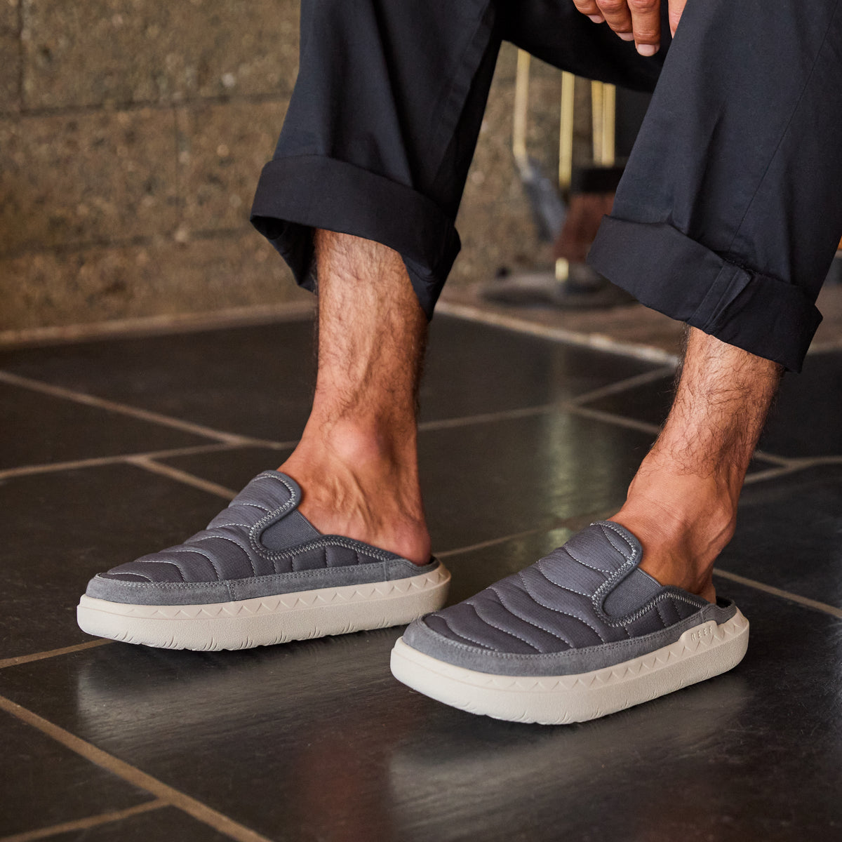Rambler Kicker slippers facing right worn indoors, featuring cushioned memory foam footbed and lightweight EVA outsole in charcoal gray