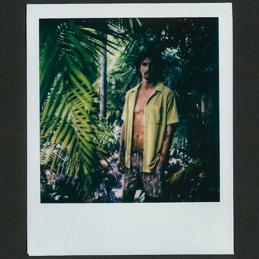 Man wearing open Myran Camp Shirt standing among lush green tropical plants, surf-inspired casual beach apparel.