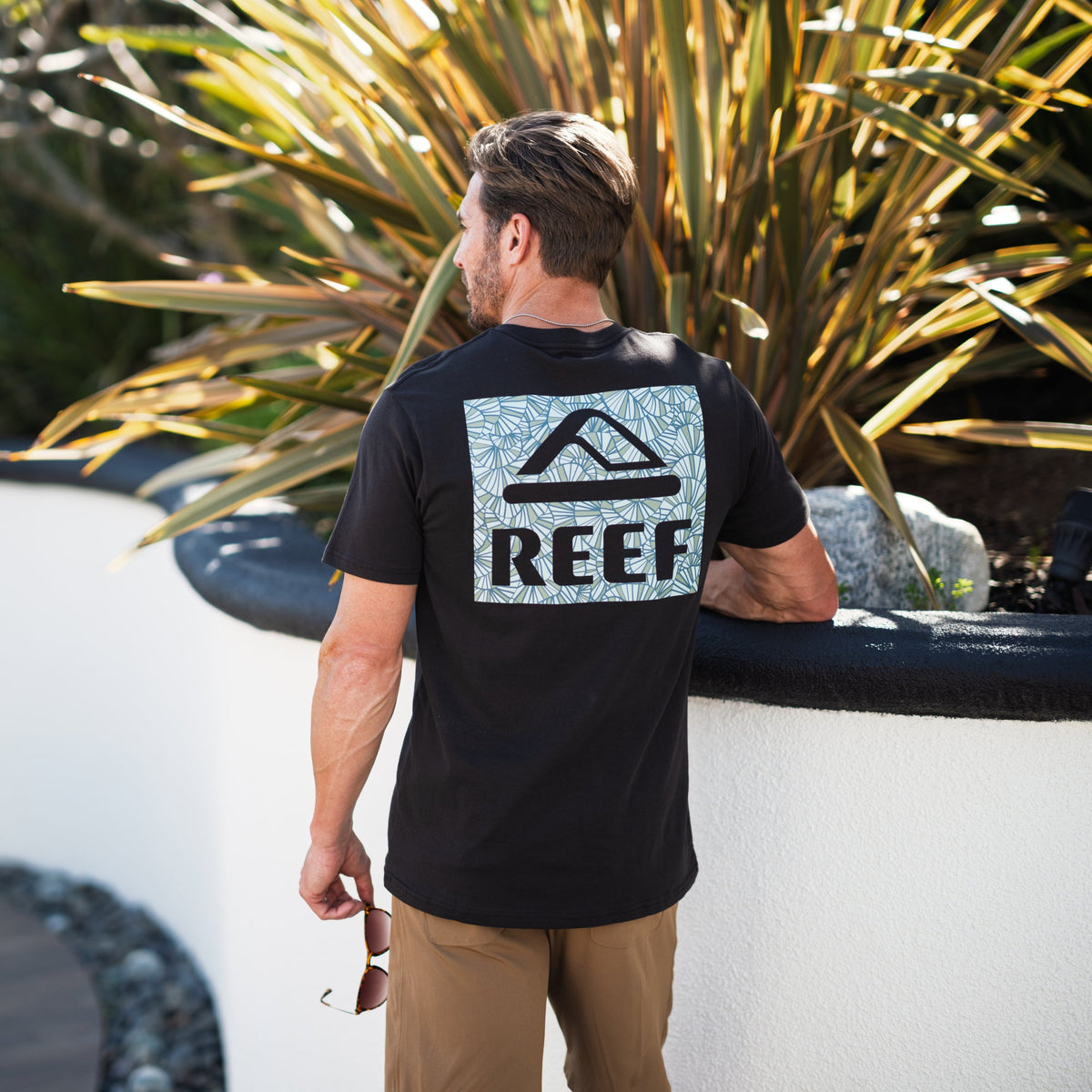 Man wearing Shells Tee facing away showing back logo, outdoors near plants, promoting beach-inspired comfort and style