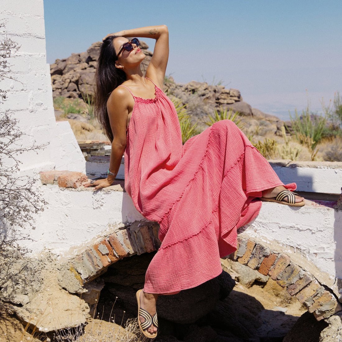 Woman wearing a coral Lainey Maxi Dress seated outdoors in sunlight, showing sandals facing forward, surf-inspired comfort style.