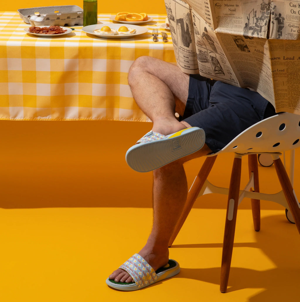 Mulligan Slide worn by man sitting, left foot facing forward showing slide top, right foot raised showing outsole.