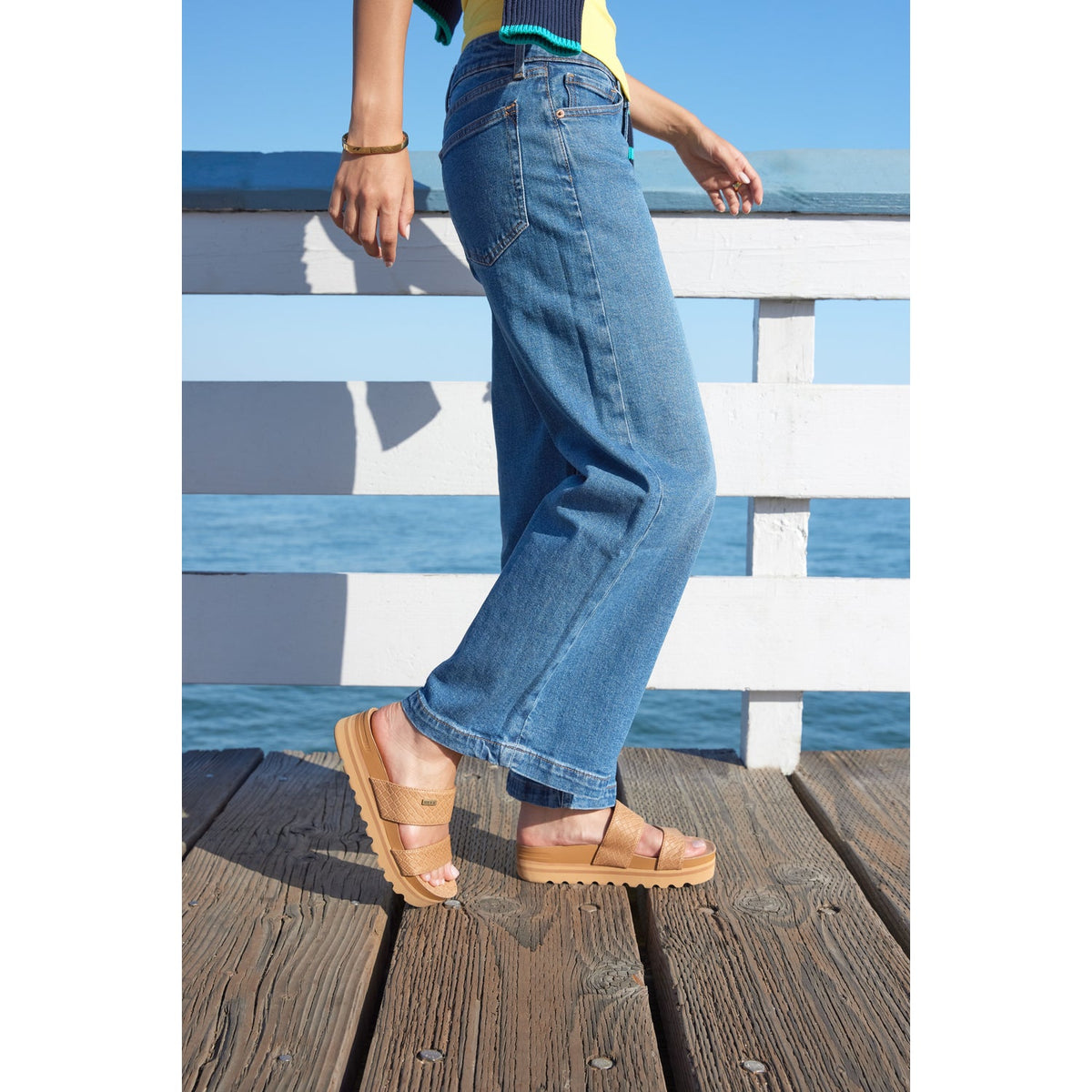Side view of women's Cushion Vista Hi tan sandals on a wooden pier, highlighting comfort and beach-inspired design.