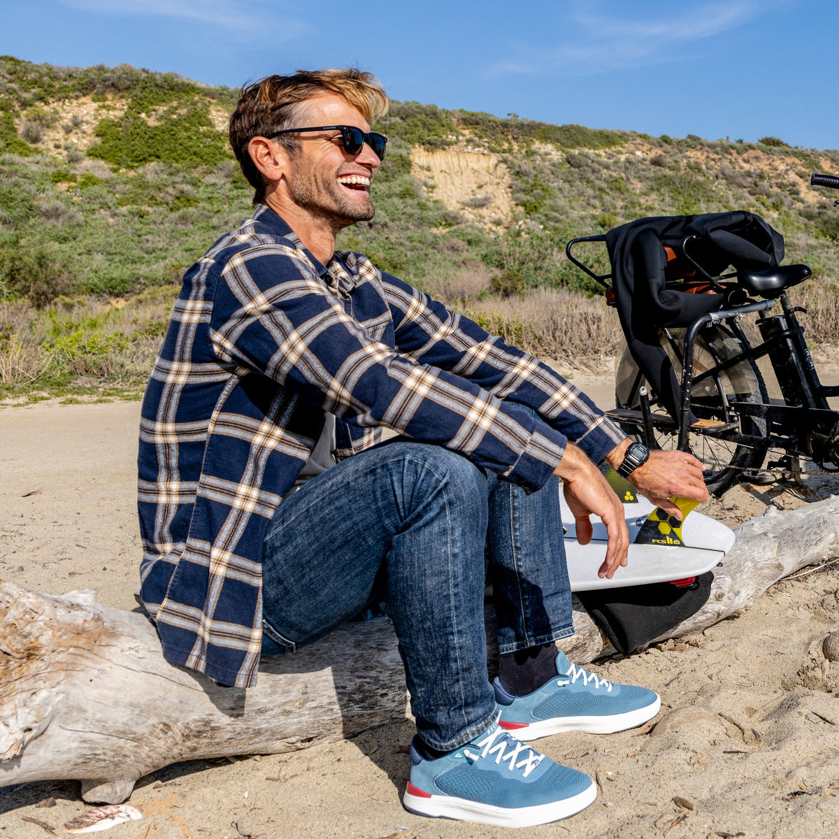 Man wearing Shoreline Del Mar shoes facing left, seated on driftwood at the beach with sunglasses and casual outfit.