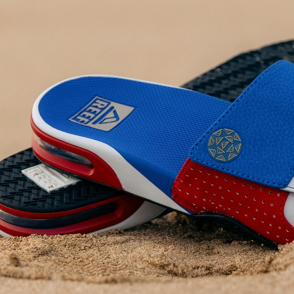 Pair of Fanning Slide sandals facing left in red, white, and blue with cushioned EVA footbeds on sand background